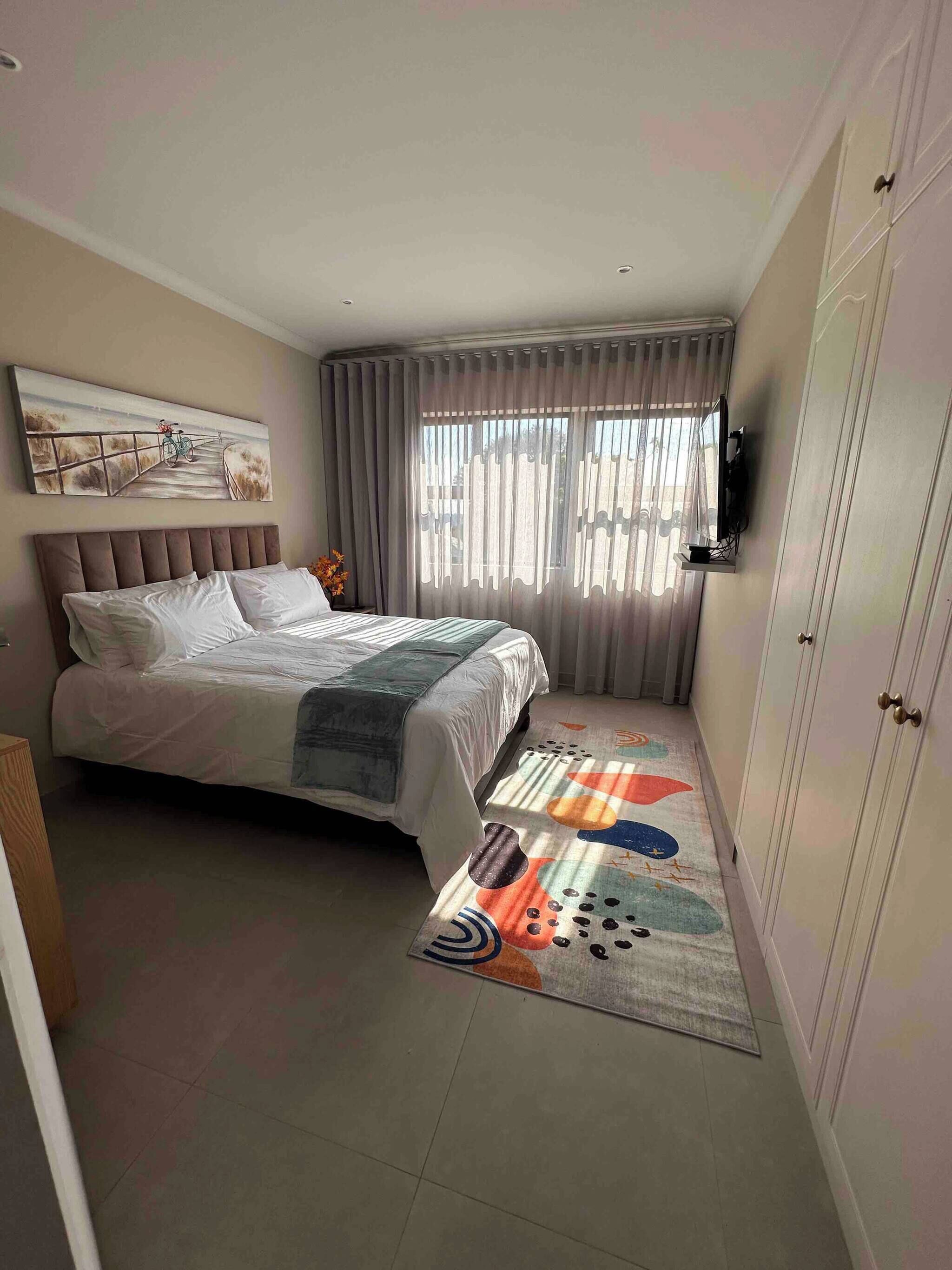 Un dormitorio con cama, televisor sobre un mueble, un cuadro en la pared y ventana con cortinas.