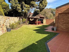 A well-maintained backyard with a stone wall, a grassy lawn, a barbecue area, and a brick building. - Kalistas House Villa (Johannesburg)