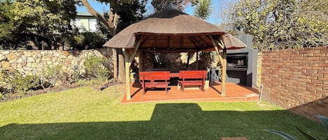 A patio with a thatched roof, two wooden benches, a grill, and a stone wall.