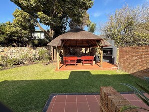A patio with a thatched roof, two wooden benches, a grill, and a stone wall. - Kalistas House Villa (Johannesburg)