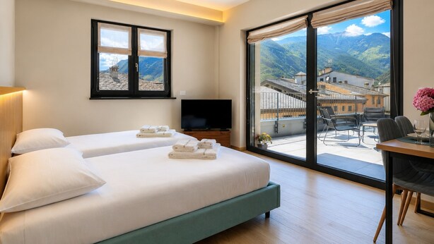 A hotel room with two beds, a TV, a balcony with a view of mountains, and a dining area.