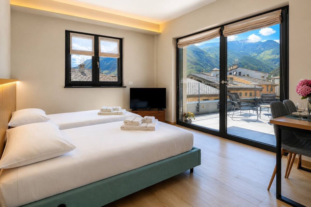 Habitación de hotel con dos camas, televisor, balcón con vistas a las montañas y un comedor.