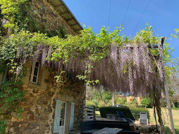 Un bâtiment en pierre doté d’un porche couvert, d’un banc en bois et d’un jardin agrémenté de verdure et d’une pergola ornée de plantes grimpantes.