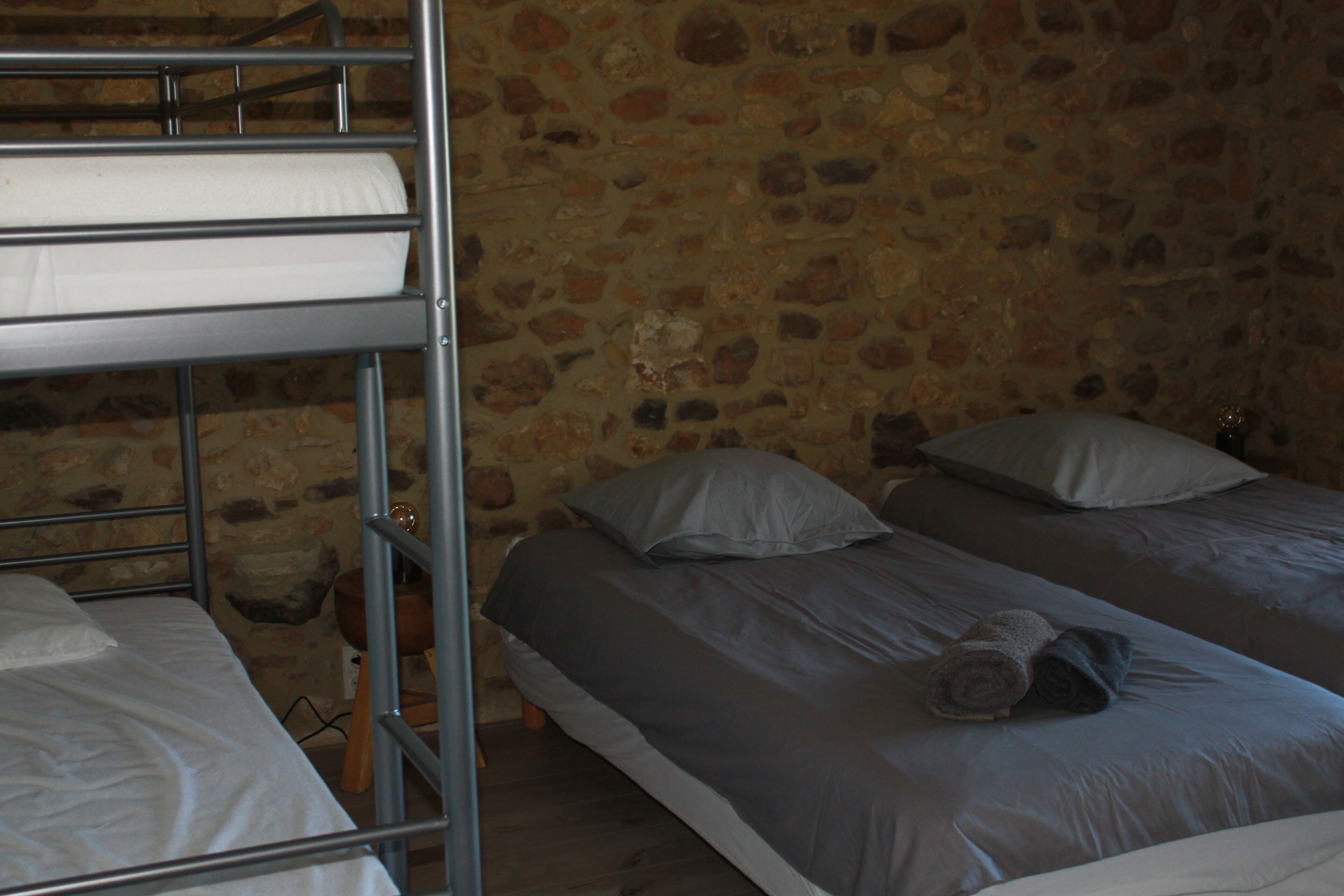Habitación con literas, pared de piedra, taburete de madera y una toalla enrollada sobre la cama.