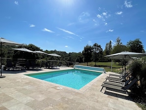 Una piscina rectangular con sillas de descanso y sombrillas, rodeada por un solárium con baldosas y vegetación.