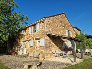 Un bâtiment en pierre avec un toit en pente, de nombreuses fenêtres avec volets, des sièges à l’extérieur et un sol recouvert de gravier.