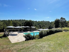Un espace piscine doté de chaises longues et de parasols, entouré d’une pelouse verdoyante et d’arbres.