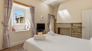 A bedroom with a bed, a television, a window with curtains, a fireplace, and a stone wall. - Ghyllstead - North East Escapes (Glanton)