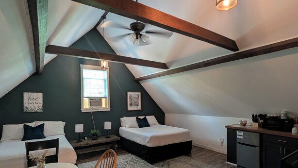 Ein Schlafzimmer im Loft-Stil mit schrägem Dach, Deckenventilator, Bett, Schreibtisch und einer Küchenzeile.