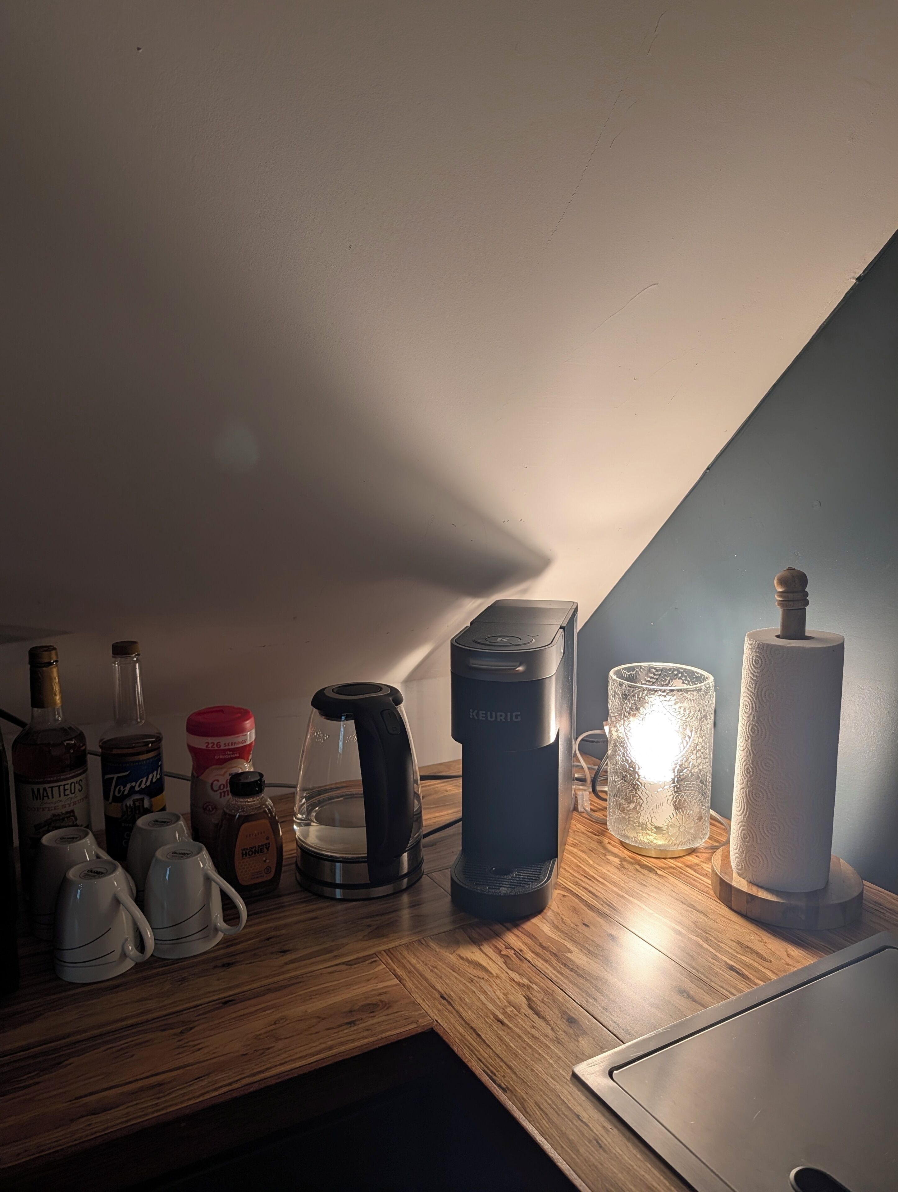 A kitchenette with a coffee maker, a glass carafe, a kettle, a jar of honey, and a roll of paper towels on a wooden countertop.