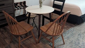 Ein kleiner, gemütlicher Raum mit einem runden Tisch, zwei Holzstühlen, einem Bett mit weißer und blauer Tagesdecke, einem Nachttisch und einem Wandregal.