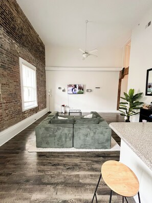 Un salon comprenant un canapé d’angle, une table basse, un téléviseur fixé au mur, un ventilateur de plafond et un mur en briques.