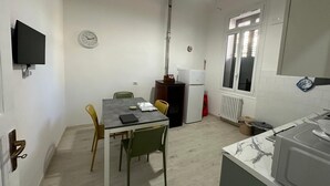 A compact kitchen with a dining area, a clock, a window with blinds, and a TV mounted on the wall. - Casa Pinuccio Sapri: Bike-Friendly Holiday Home Steps from the Sea (Sapri)