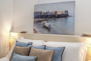 A bedroom with a bed, pillows, a wall-mounted painting of a coastal scene, and bedside lamps. - Lapuritate Via Cariddi - Bilo terrazza sul mare (Gallipoli)