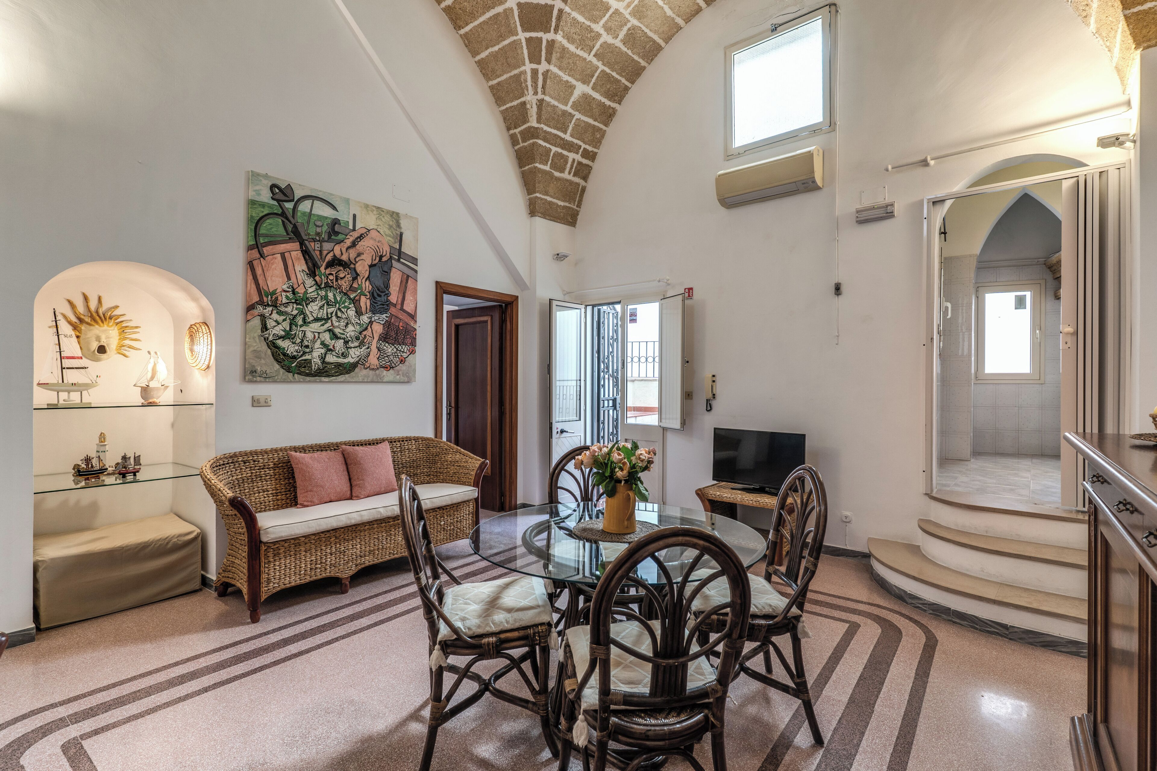 A spacious living area with a round glass table, wicker furniture, and a large painting on the wall.