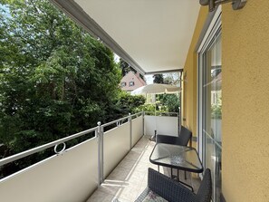 Uma varanda com mesa de vidro e duas cadeiras, um guarda-sol branco e vista para árvores e uma casa.