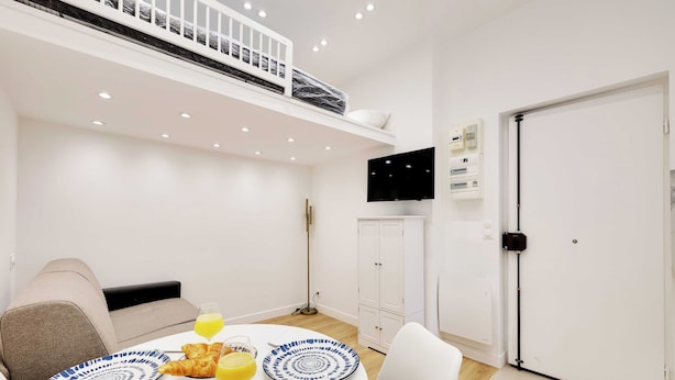 Una habitación moderna con una litera, una mesa de comedor con platos y croissants, un sofá, un televisor y una puerta.