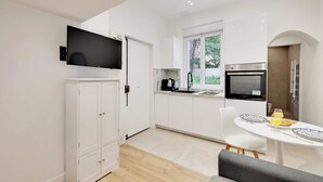 Una cocina moderna con armarios blancos, horno empotrado, televisor y comedor con mesa y sillas.
