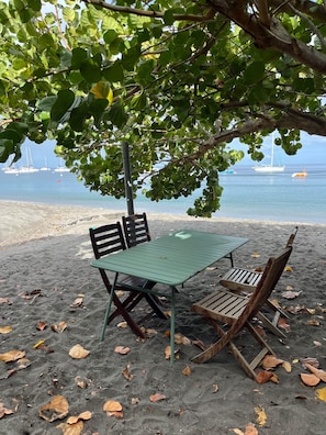 A beach with a table and chairs under a tree, a sandy shore, and boats in the water. - The House on the Beach (Saint-Pierre)