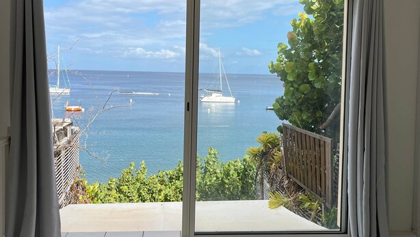 A room with a large sliding glass door leading to a balcony overlooking a marina with boats and a clear sky. - The House on the Beach (Saint-Pierre)
