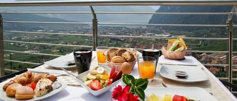 A breakfast table with croissants, fruit, and juice, overlooking a mountainous landscape.