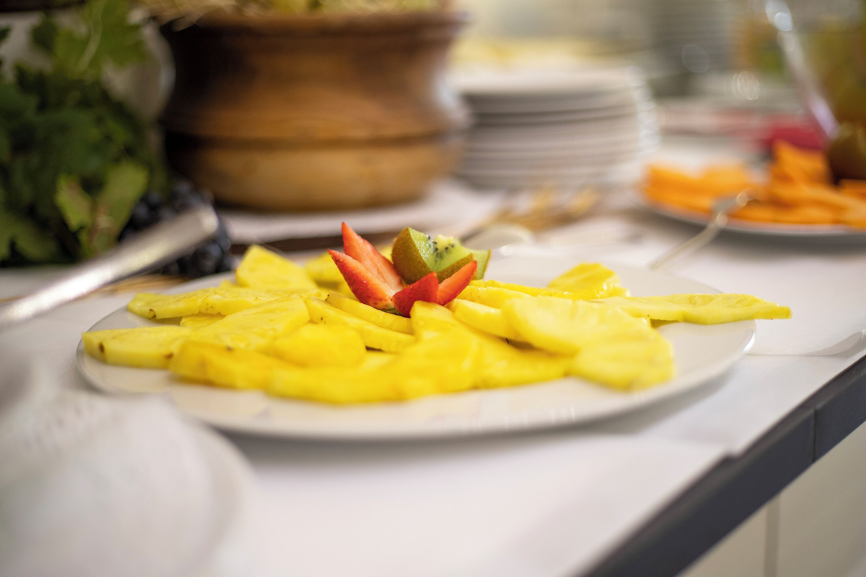 Une table de buffet avec une assiette de fruits jaunes disposés en éventail, un saladier de salade verte et une pile d’assiettes en arrière-plan.