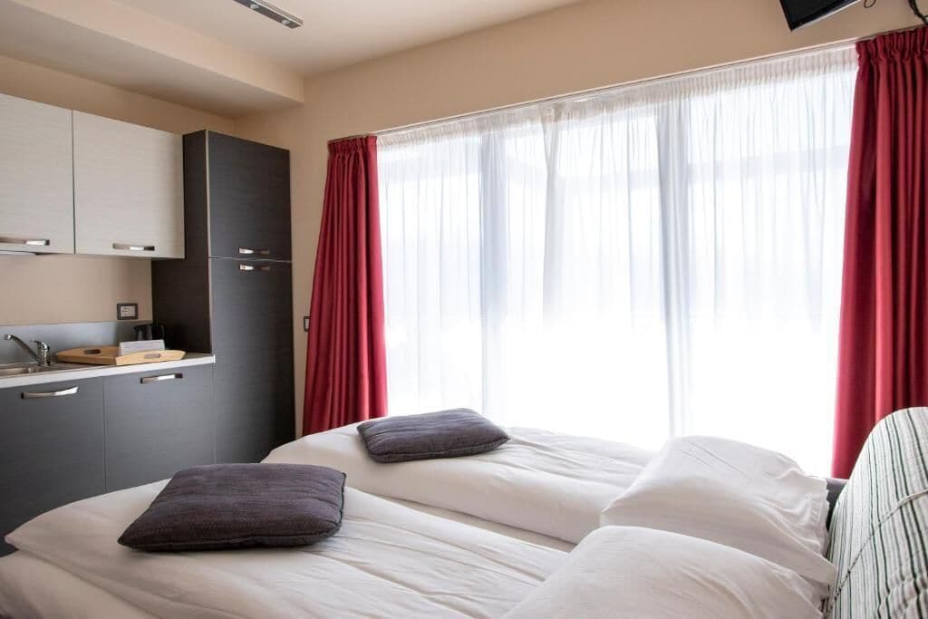 A hotel room with a bed, a TV, a kitchenette, and a window with curtains.