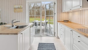 A modern kitchen with white cabinets, a wooden countertop, a stainless steel sink, and a glass door leading to an outdoor patio. - 5 Star Holiday Home in Vig (Vig)