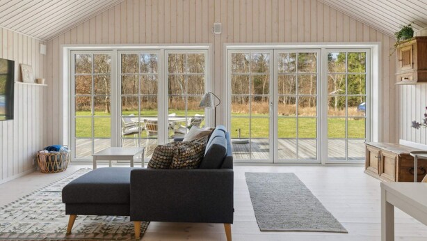 A spacious room with a high ceiling, a large sliding glass door leading to an outdoor area, a dark grey sofa, a small table, and a lamp. - 5 Star Holiday Home in Vig (Vig)