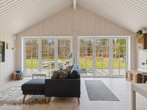 Ein geräumiger Raum mit hoher Decke, einer großen Schiebeglasdoor zum Außenbereich, einem dunkelgrauen Sofa, einem kleinen Tisch und einer Lampe.
