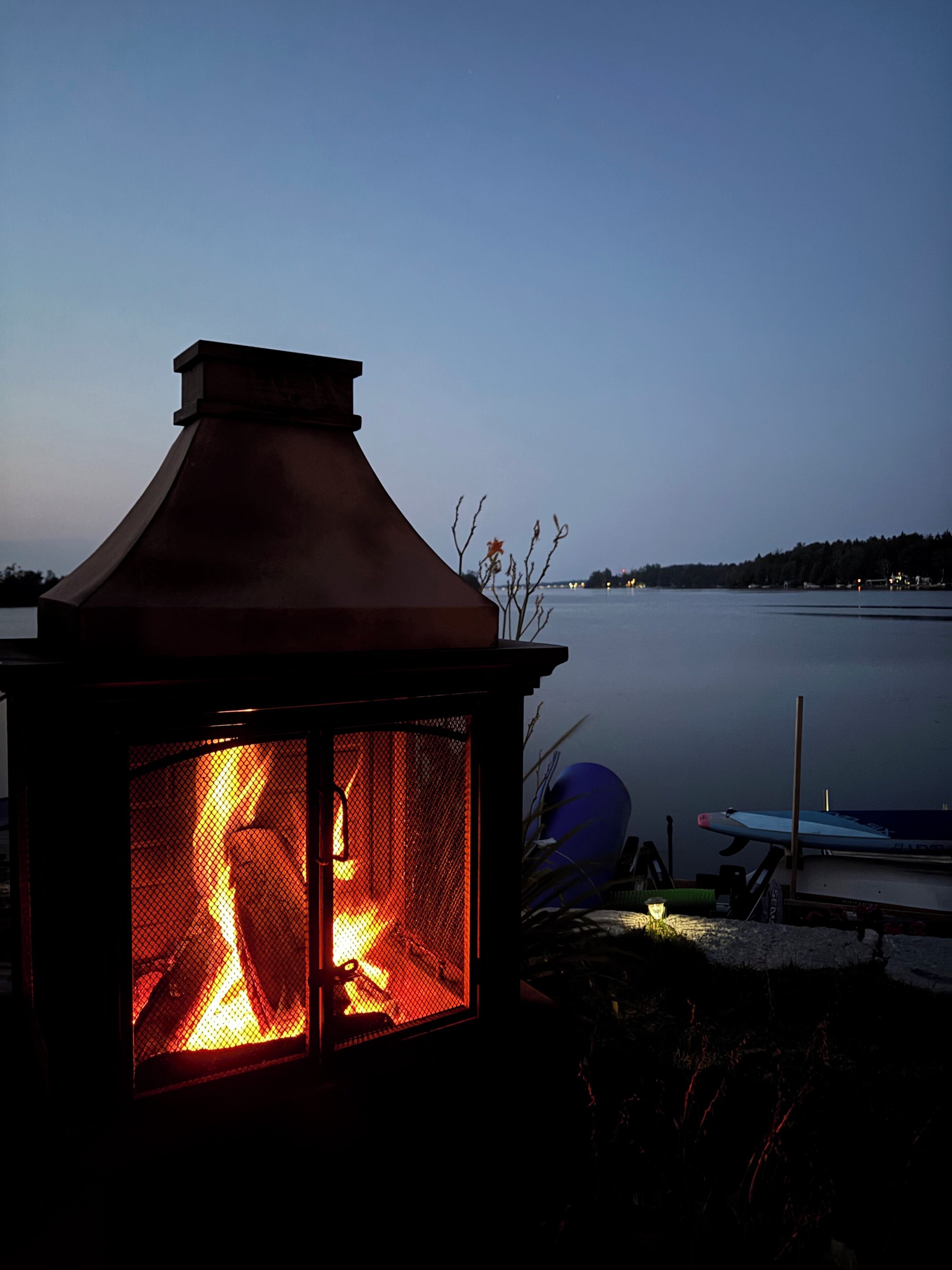 A fire within a metal lantern structure, overlooking a calm body of water with boats docked.
