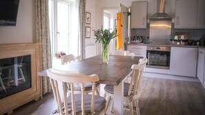 Un coin repas avec une table en bois, des chaises blanches, un vase de fleurs et une cuisine en arrière-plan.