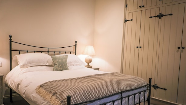 Una cama con estructura metálica, ropa de cama blanca, un plaid estampado, una almohada verde, una lámpara de noche y un armario con tiradores verticales.