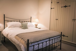 Ein Bett mit Metallgestell, weißer Bettwäsche, einer gemusterten Tagesdecke, einem grünen Kissen, einer Nachttischlampe und einem Kleiderschrank mit vertikalen Griffen.