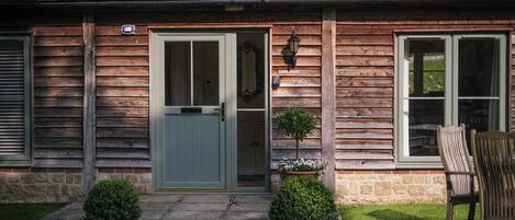 A wooden cottage with a front door, two windows, a small porch, and a chair outside.