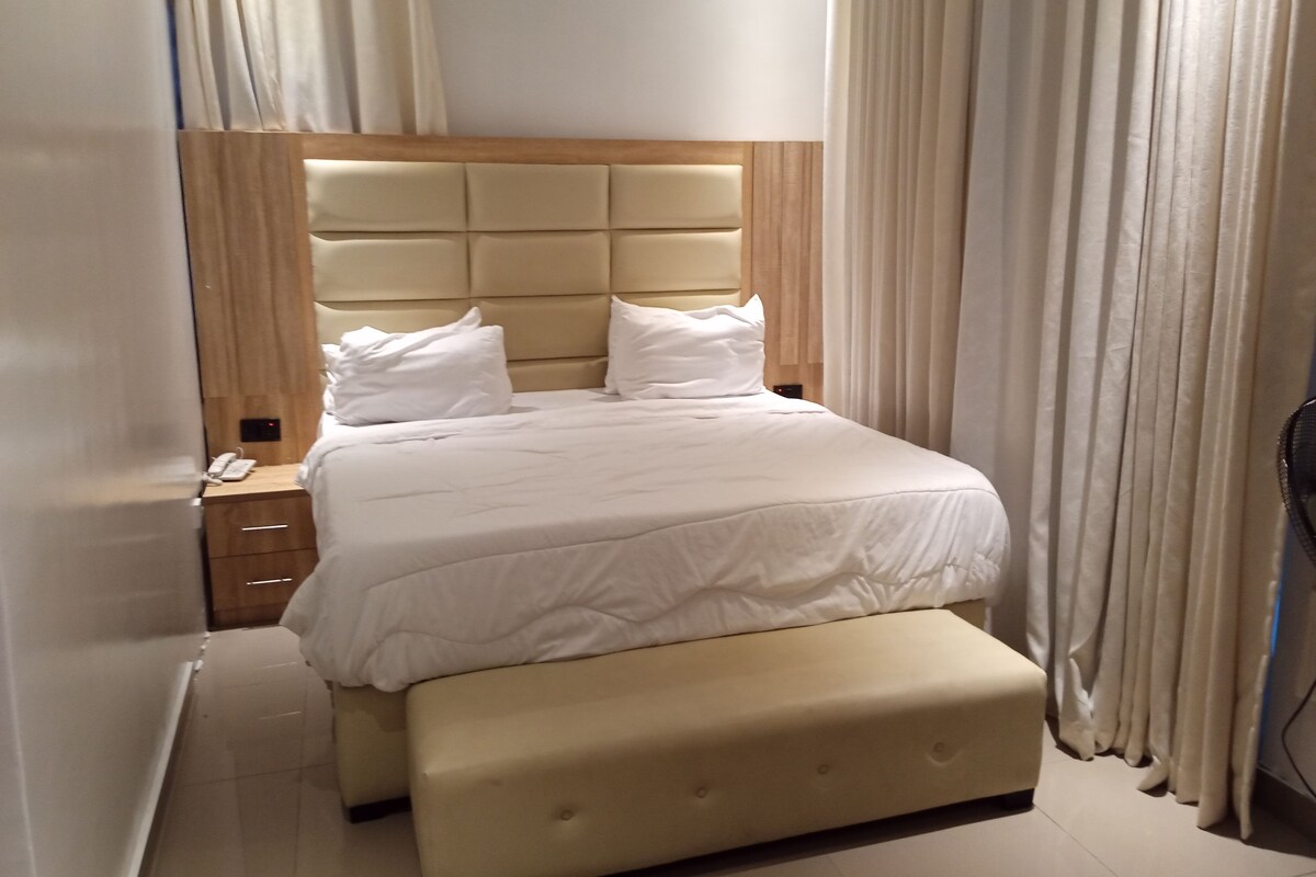 Una habitación de hotel con cama, mesita de noche, lámpara y ventana con cortinas.