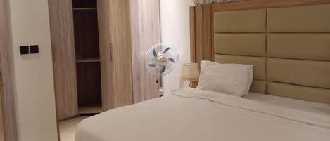A bedroom with a bed, a bedside lamp, a wall-mounted fan, and a wardrobe.