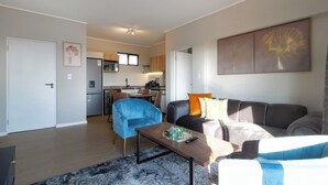 A modern living room with a sofa, a blue armchair, and a coffee table. There is a kitchen in the background with wooden cabinets and a dining area. - Captain's Akaru Hide Away (Sandton)