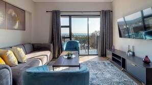 A modern living room with a sofa, armchair, and coffee table, a balcony with curtains, a TV, and a city view. - Captain's Akaru Hide Away (Sandton)