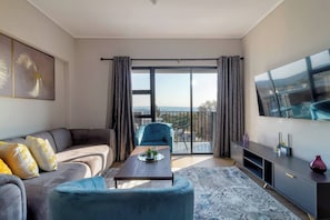 Ein modernes Wohnzimmer mit Sofa, Sessel und Couchtisch, ein Balkon mit Vorhängen, einem Fernseher und Blick auf die Stadt.