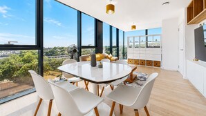 A modern dining room with a large table, white chairs, and a city view through large windows. - Bright 1BR Home with Shared Terrace-1min to Tube (London)