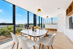 A modern dining room with a large table, white chairs, and a city view through large windows. - Bright 1BR Home with Shared Terrace-1min to Tube (London)