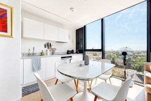 A modern kitchen with white cabinets, a glass dining table, and a view of the city through large windows. - Bright 1BR Home with Shared Terrace-1min to Tube (London)