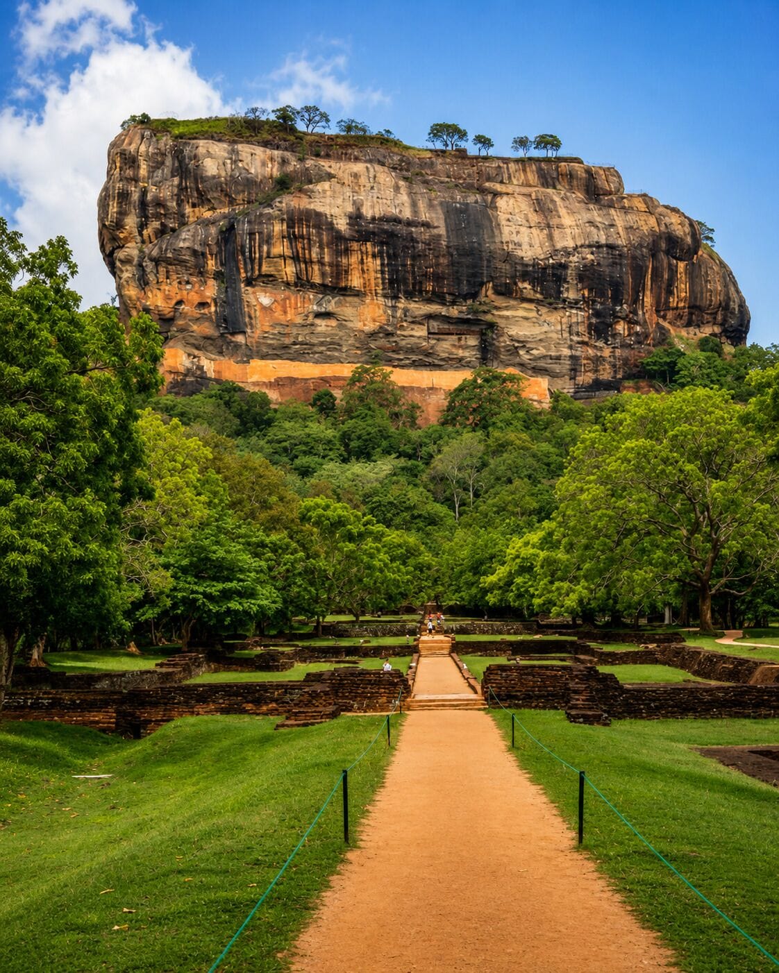 A pathway leading to a large rock formation with trees and ruins in the foreground.