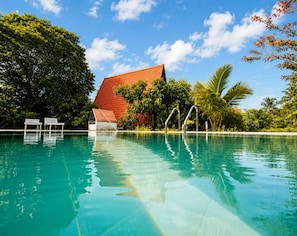 A clear swimming pool with a red-roofed building in the background, surrounded by lush greenery.
