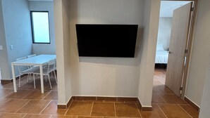 Ein Zimmer mit einem an der Wand befestigten Flachbildfernseher, einem Esstisch mit Stühlen und einem gefliesten Boden.