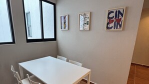 Ein Zimmer mit einem weißen Tisch, Stühlen, zwei Fenstern und drei gerahmten Kunstwerken an der Wand.