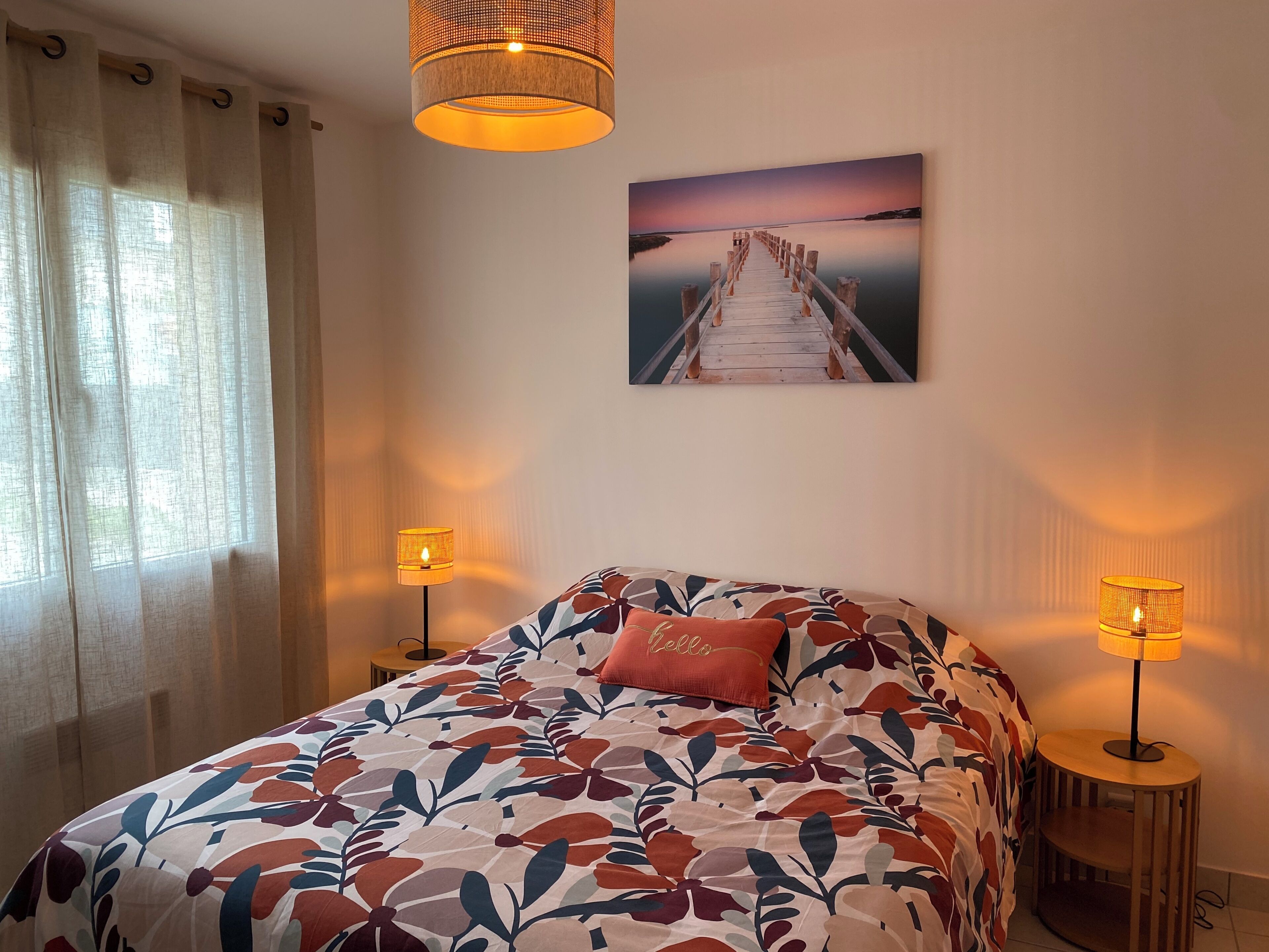 A bedroom with a bed, two bedside lamps, a window with curtains, a wall-mounted picture, and a small round table.