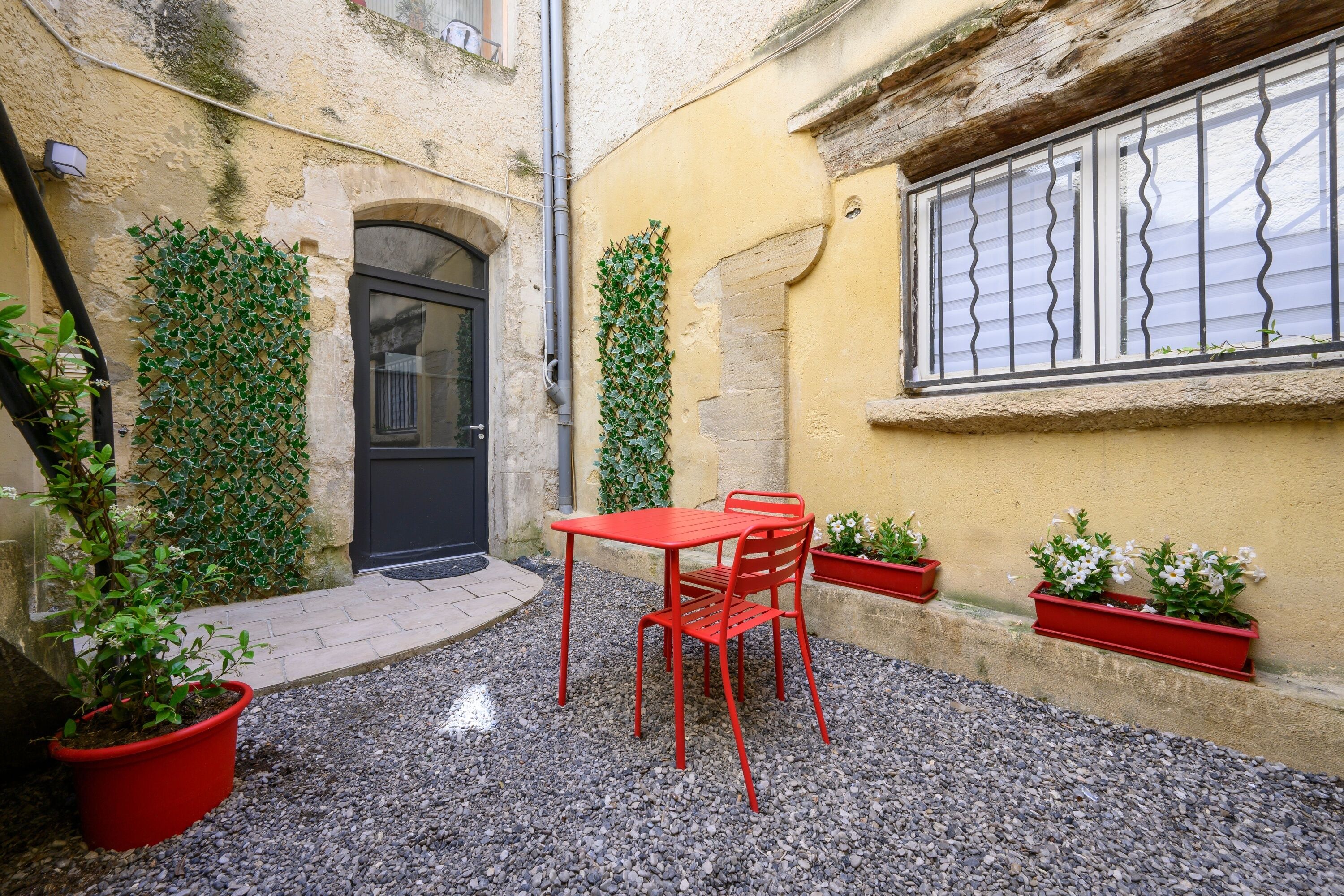 Une cour avec une table et des chaises rouges, une porte noire et des plantes en pot.