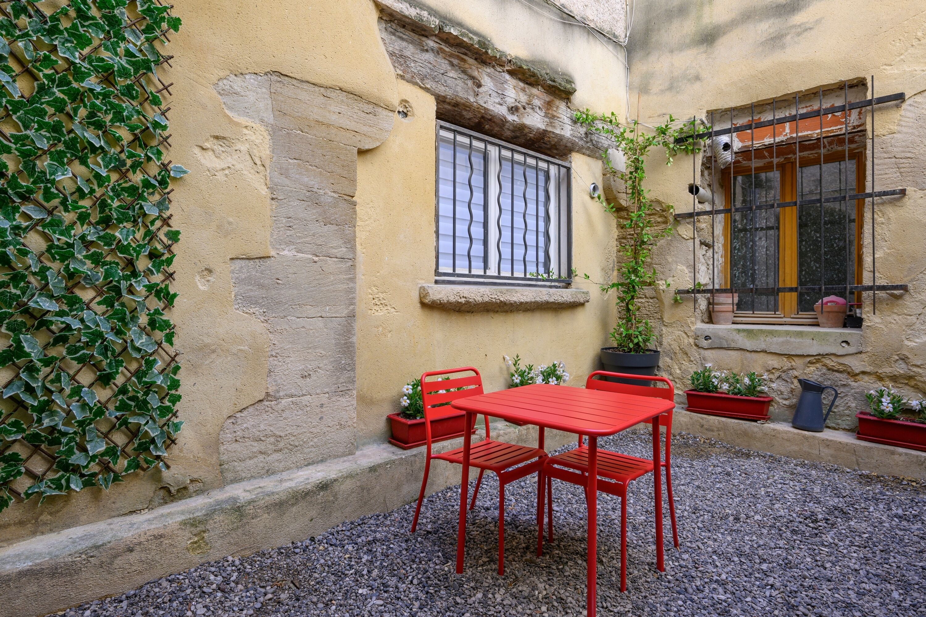 Un patio con una mesa y sillas de metal rojo, plantas en macetas y una ventana con vidrios protegidos.